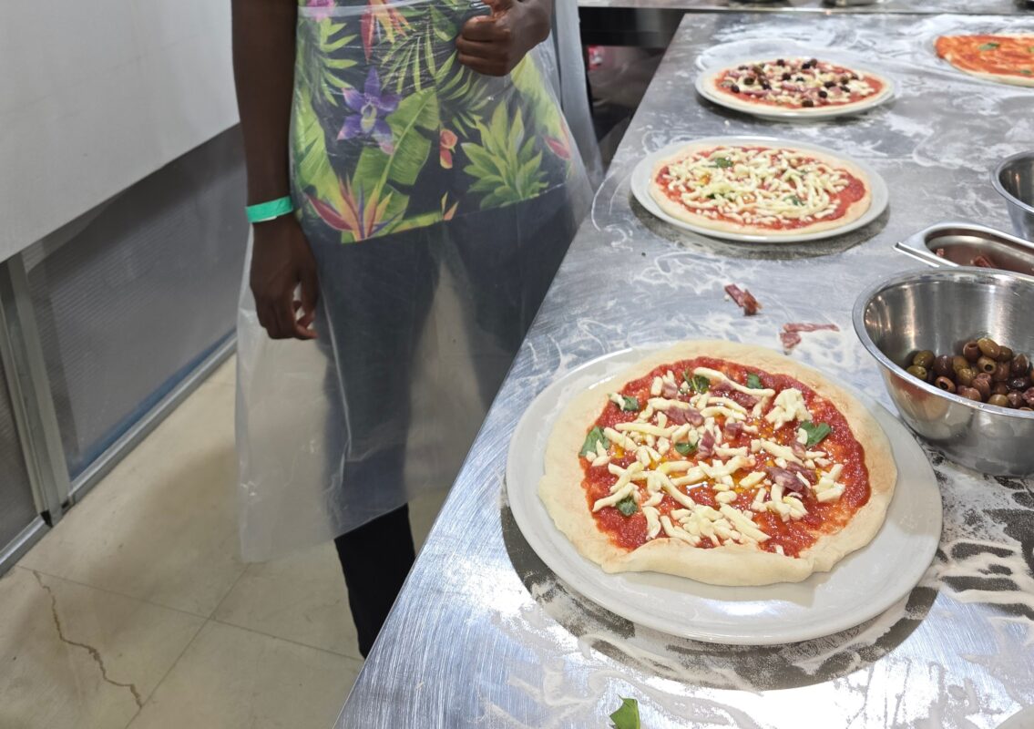 Close-up of three raw pizzas ready to be baked, surrounded by bowls of toppings and a person in an apron standing nearby.