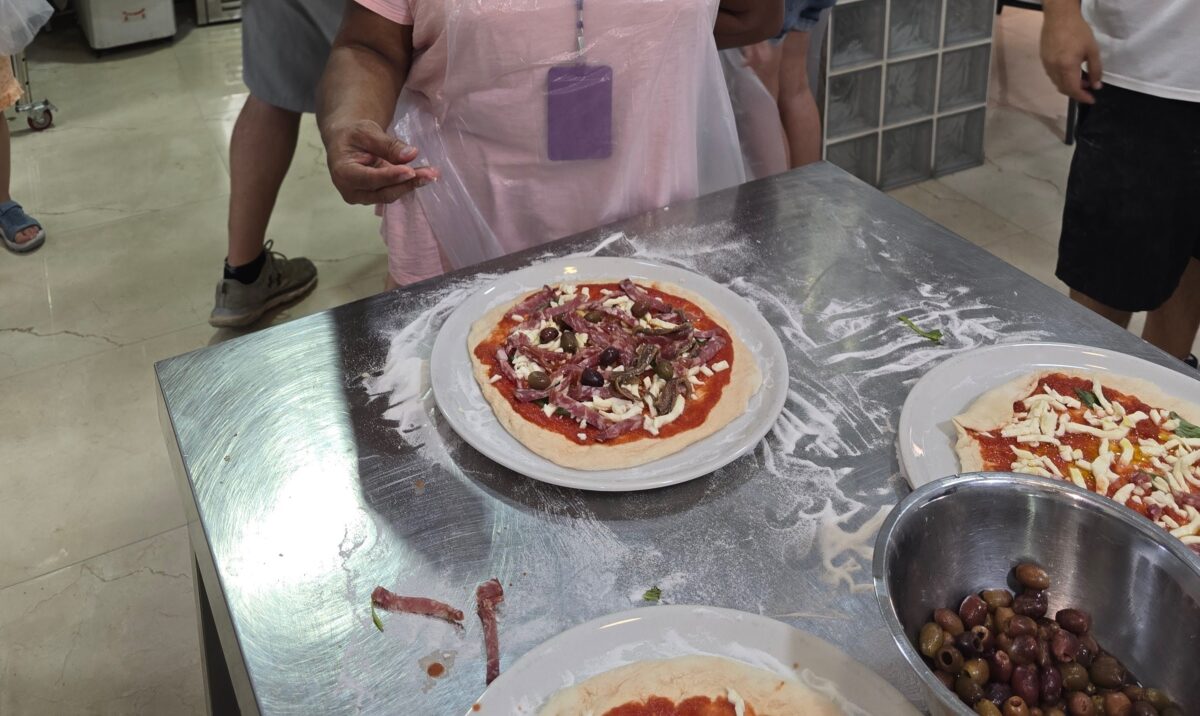 Close-up of a participant, wearing plastic apron, preparing her creation with toppings during pizza-making in Naples.