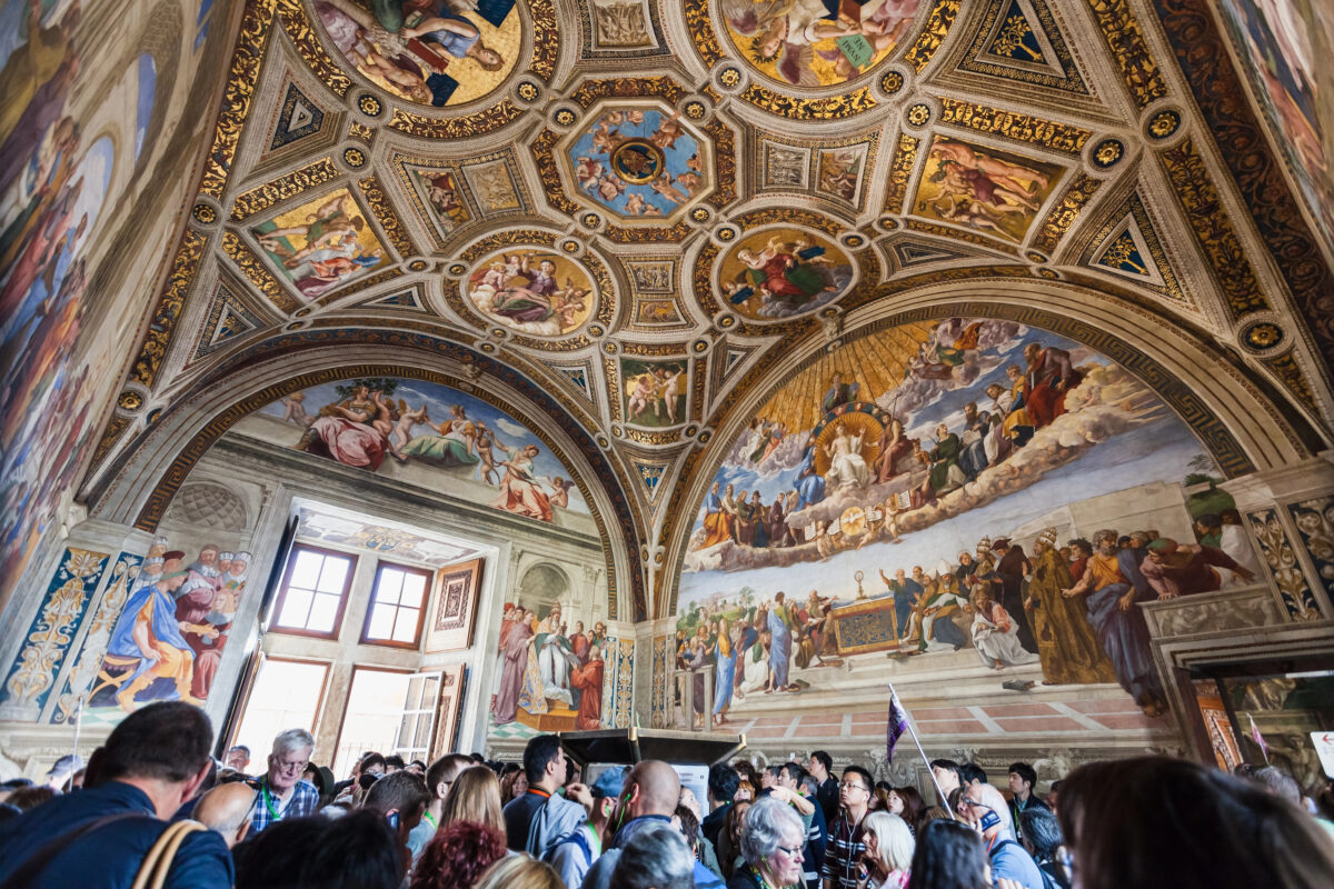 Interior of the Vatican Museums showing Raphael’s frescoes and ornate ceiling with tourists gathered below.
