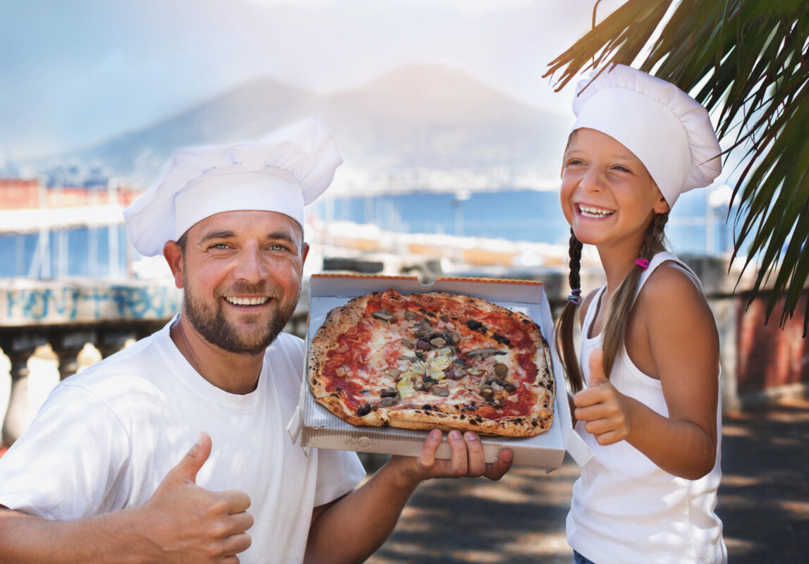 A smiling man and girl in white chef hats holding a pizza topped with vegetables and cheese, with Mount Vesuvius and the Bay of Naples in the background.