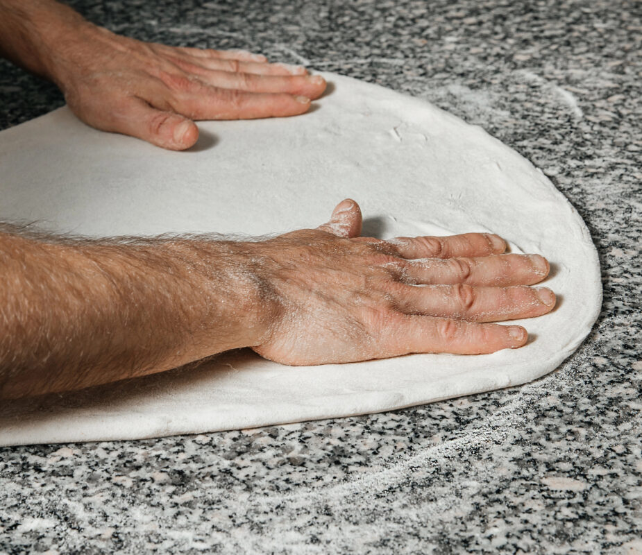 Close-up of hands pressing and stretching pizza dough on a floured granite countertop.