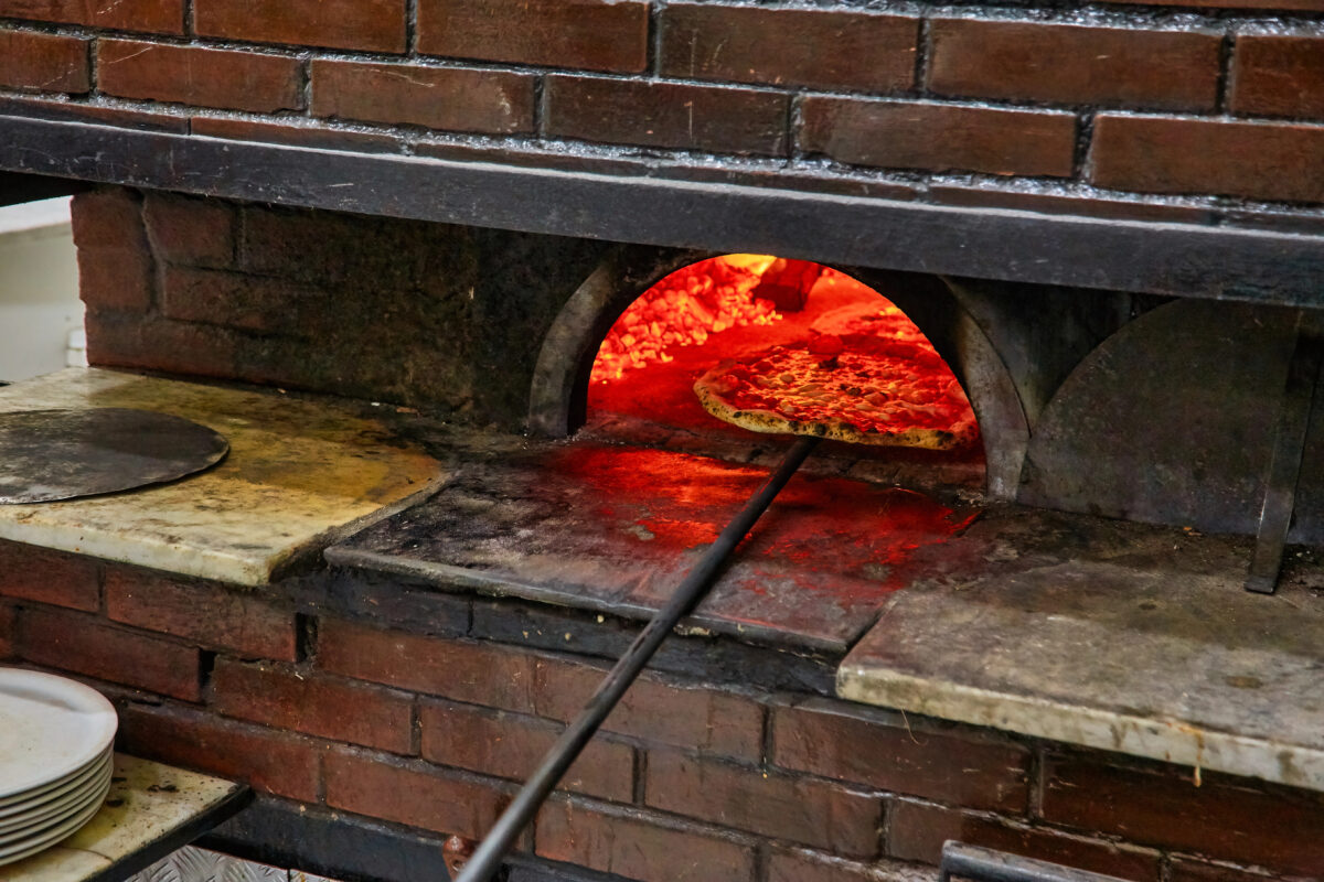 A rustic stone oven with red coals and flames visible, baking a pizza on the hot surface.