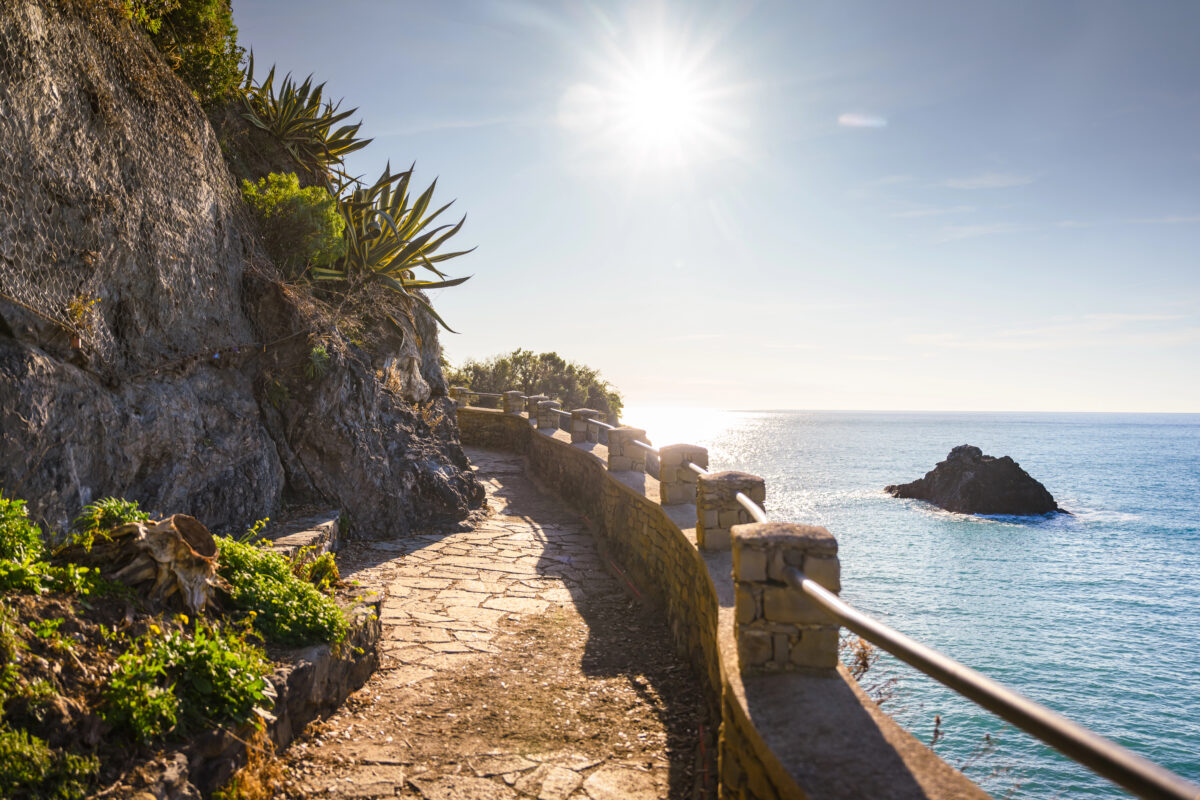 Scenic coastal path overlooking the water with bright sunlight and a rock rising from the sea.