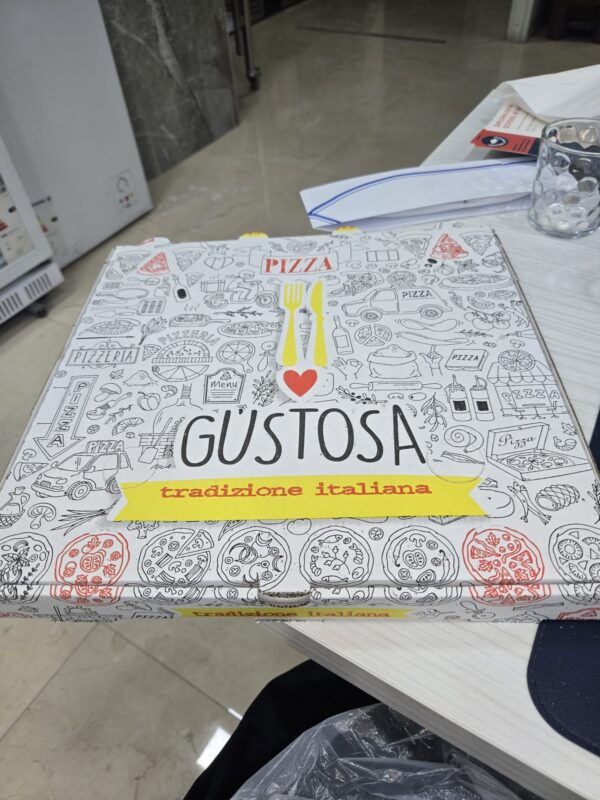 Italian pizza box with red, yellow, and black designs, sitting on a table with a glass and folded paper hat nearby.