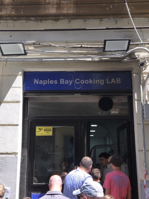 Exterior of Naples Bay Cooking Lab with a group of visitors and a chef visible inside.