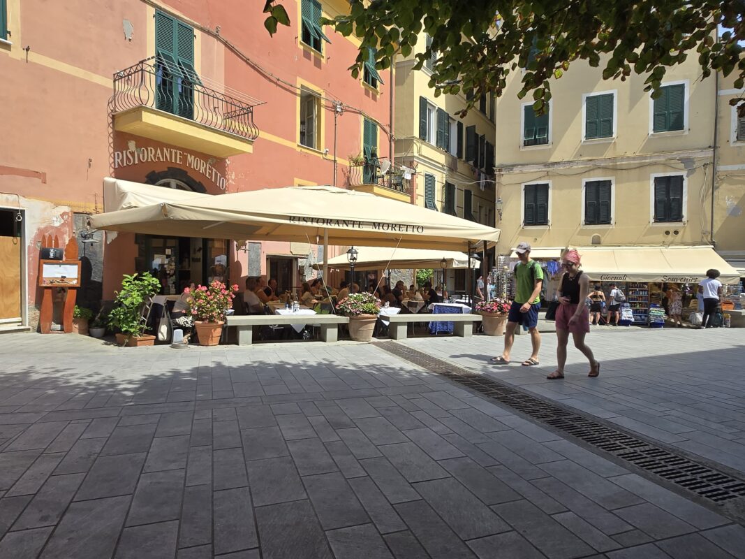 A sunny piazza in Monterosso with café seating, pedestrians, and souvenir shops nearby.