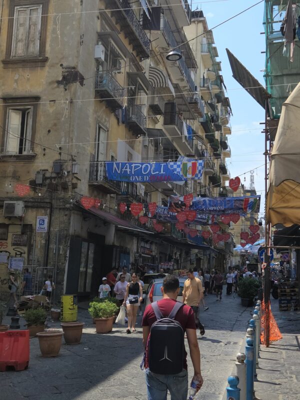 Narrow Naples street lined with balconies and laundry, decorated with banners and red heart cutouts, crowded with pedestrians.
