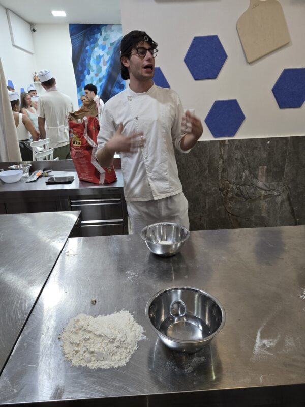 A cooking instructor in a white uniform and glasses stands behind a stainless steel counter with bowls and flour, explaining pizza-making to a class.