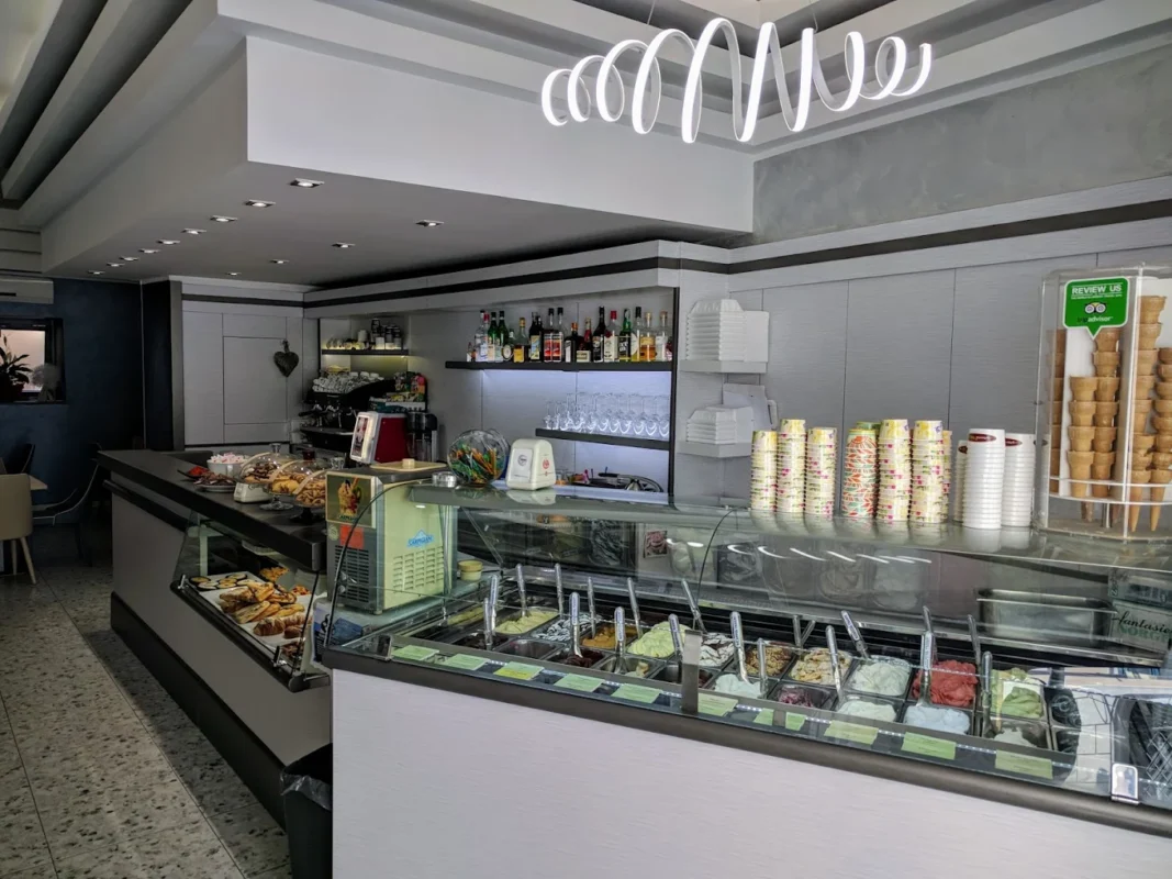 A bright, modern gelato shop with trays of colorful ice cream, pastries on the side counter, and bottles of drinks lining the shelves.