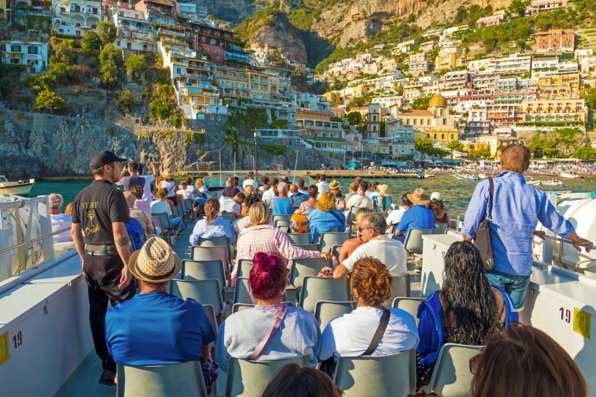 Group of cruise passengers seated on a boat with views of Positano’s stacked houses rising on a cliffside.