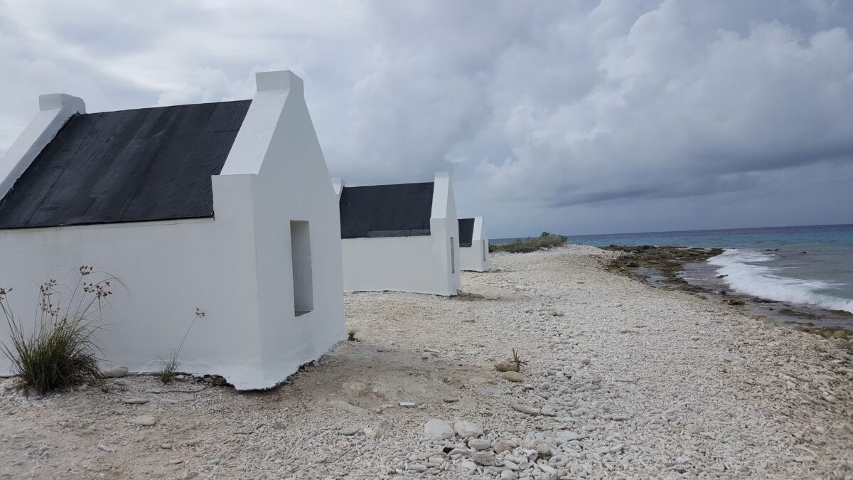 White stone slave huts on a coral-strewn beach in Bonaire, leading toward the turquoise sea beneath an overcast Caribbean sky.