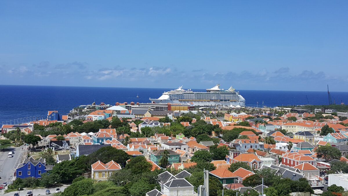 Panoramic view of Willemstad, Curaçao, showing rows of colorful Dutch colonial houses with orange roofs and a large cruise ship docked at the harbor under a clear blue sky.