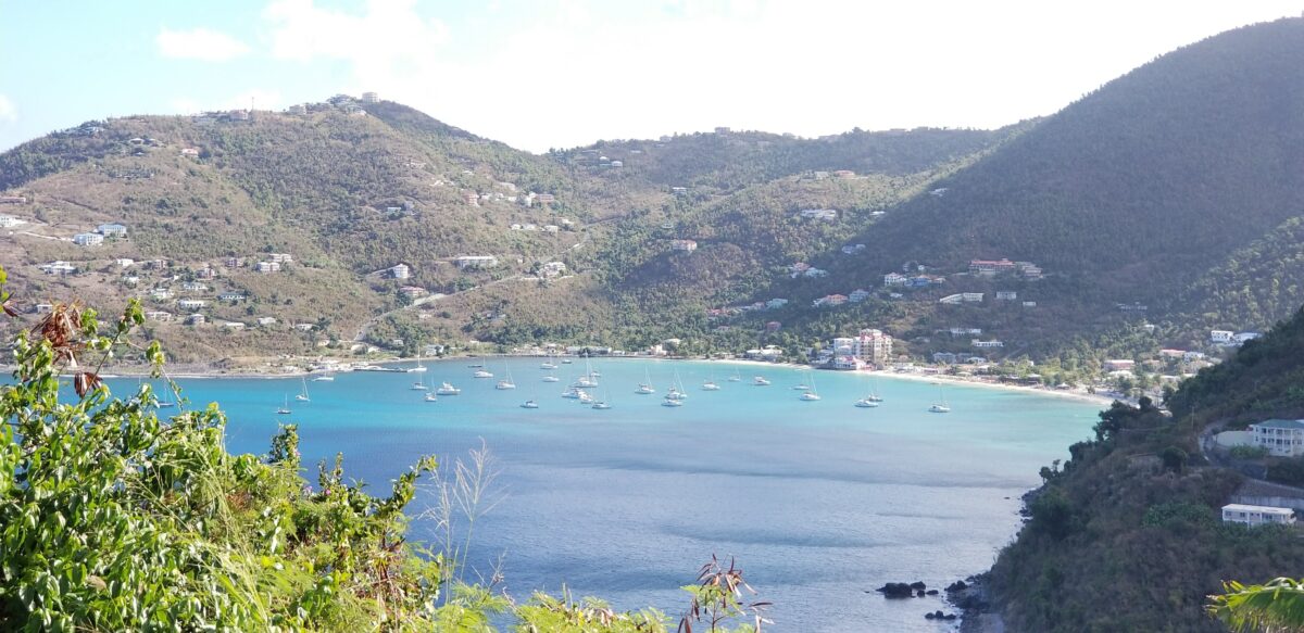 A view from a hillside on Tortola showing a calm turquoise bay dotted with sailboats, surrounded by green hills with scattered homes under a partly cloudy sky.