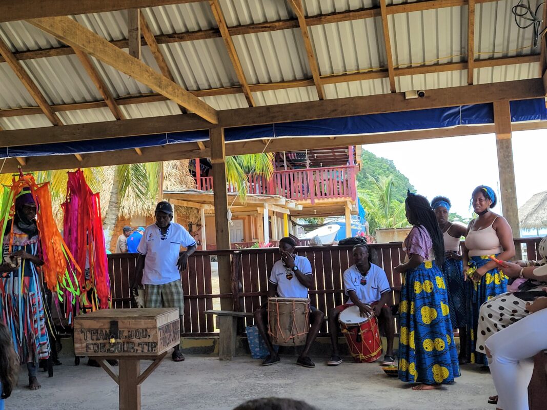 Group of Garifuna musicians and dancers performing under a covered open-air pavilion with colorful skirts, drums, and palm trees in the background.