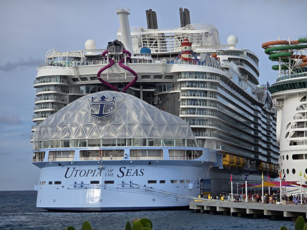 The new Utopia of the Seas moored at a Caribbean pier with its slide tower and multi-deck balconies visible against a light blue sky.
