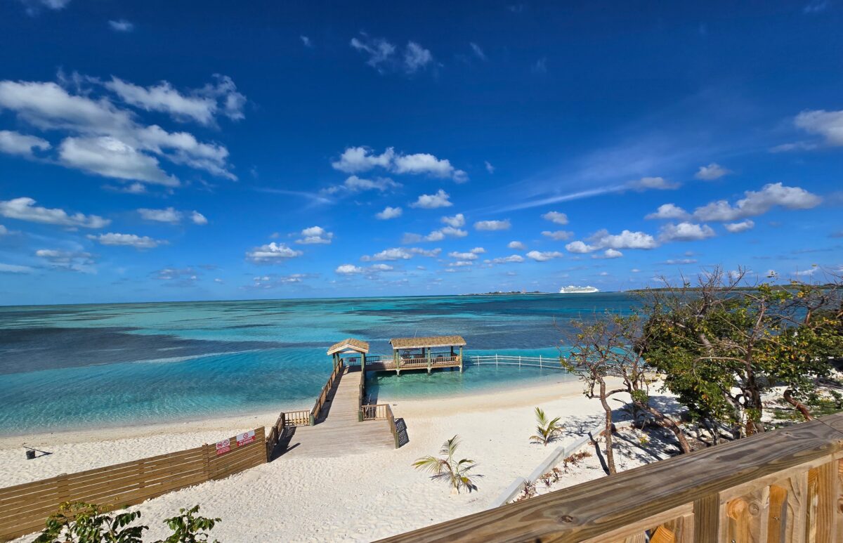 Wooden pier with small shaded pavilions extending into turquoise Bahamian water, white-sand beach and a cruise ship visible on the horizon.