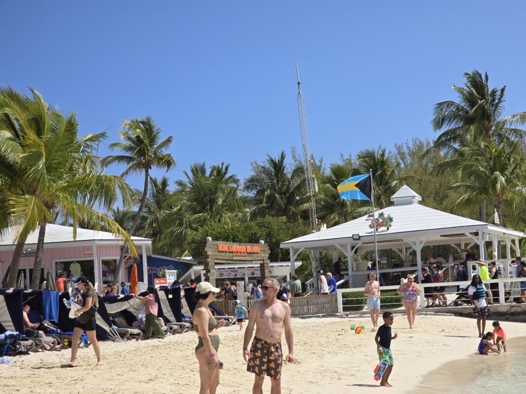 Visitors enjoying the beach at Blue Lagoon Island, Bahamas, with palm trees, cabanas, and a Bahamian flag flying near a beach bar.