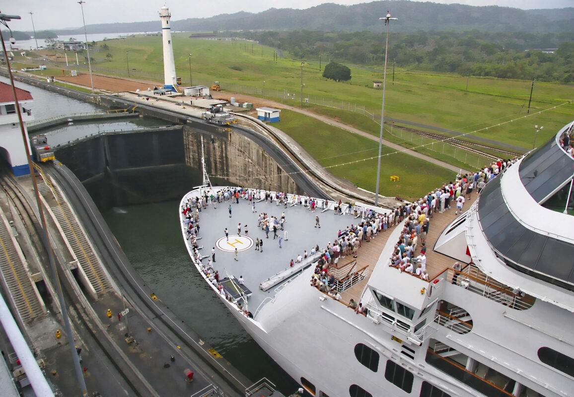 Cruise passengers standing on the bow deck as their ship passes through a lock in the Panama Canal surrounded by green hills.