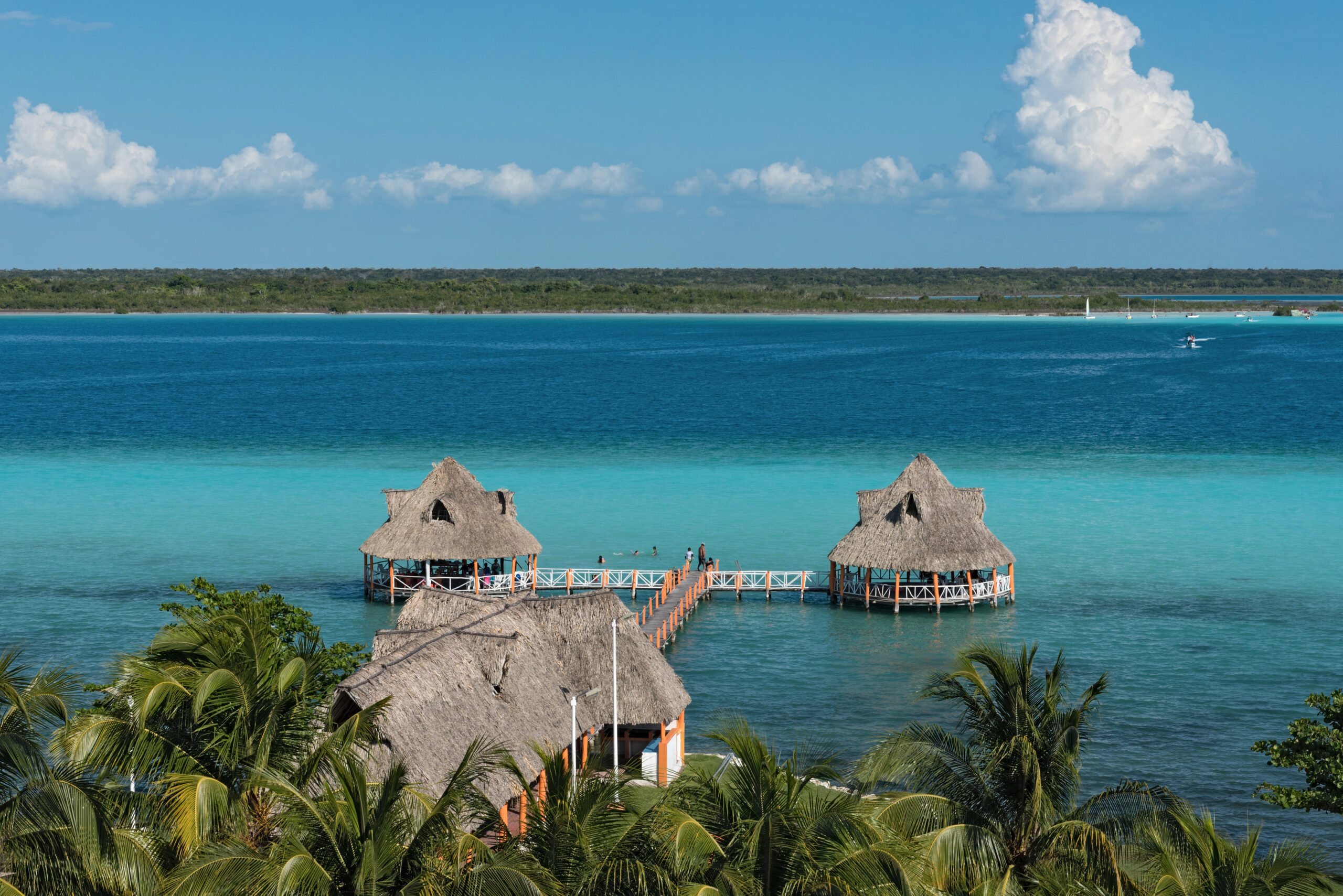 Aerial view of thatched-roof palapas connected by a wooden pier over vivid turquoise and deep blue water in Costa Maya, Mexico.