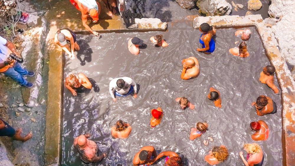 Aerial view of travelers soaking in rectangular pools of warm gray volcanic mud surrounded by stone walls at Sulphur Springs in St. Lucia.
