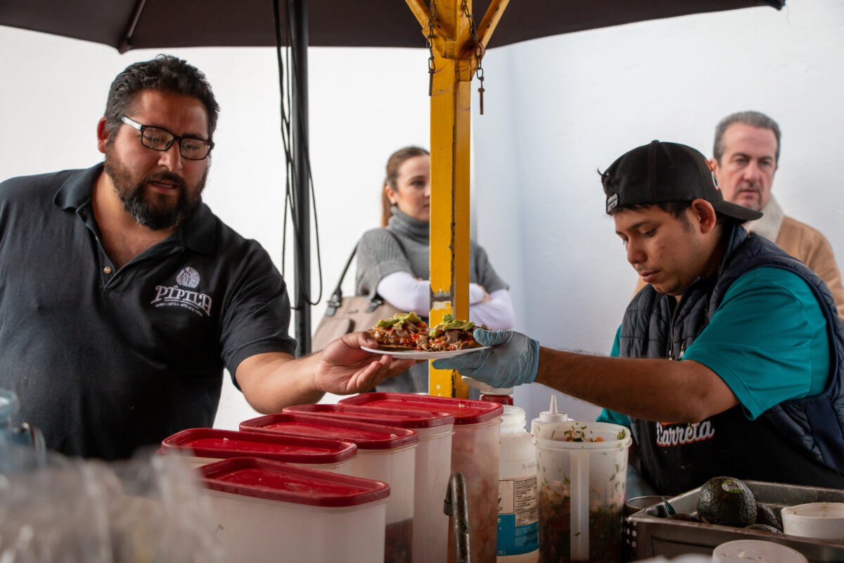 Vendor wearing gloves assembling seafood dishes at an Ensenada street stall while customers wait nearby.