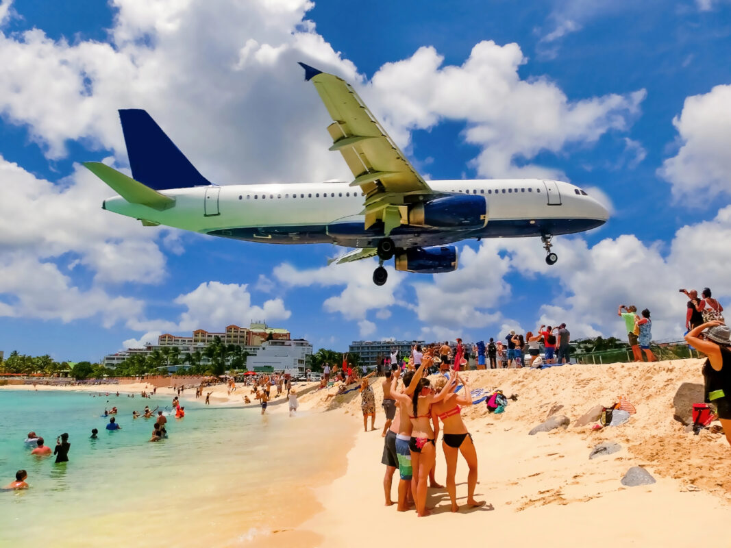 Jet flying low over a crowded beach in St. Maarten, with people in swimsuits watching and photographing as the plane prepares to land against a bright blue sky.