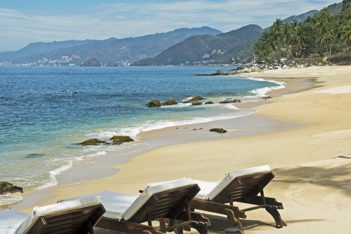 Beach chairs facing calm blue water at Playa Las Gemelas near Puerto Vallarta, with mountains and palm trees in the background.