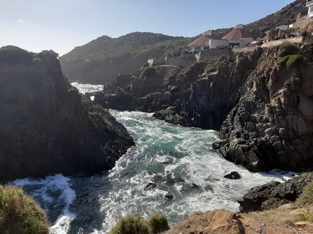 View of La Bufadora blowhole near Ensenada, Mexico, with waves surging between rocky cliffs under a clear blue sky.