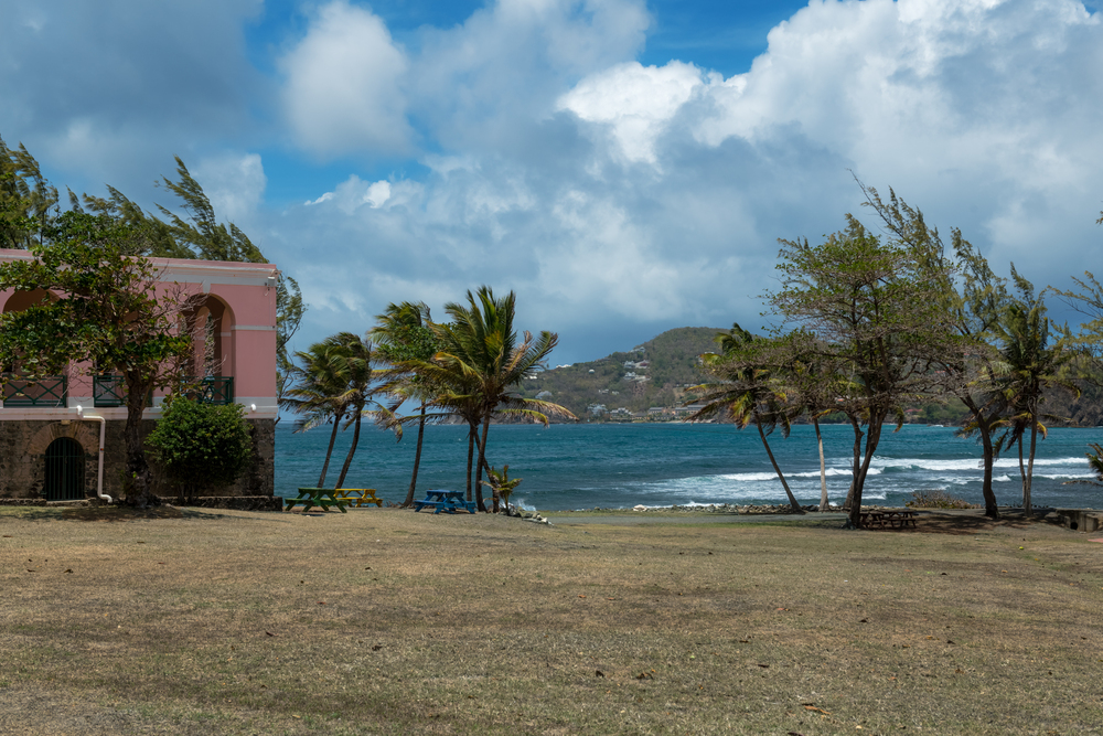 View of grassy open land with palm trees and a pink colonial-style building near the turquoise Caribbean shoreline on St. Lucia.