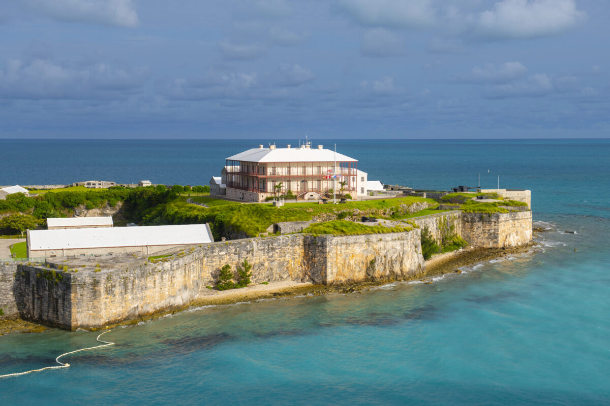 Aerial view of the Commissioner’s House in Bermuda surrounded by grassy ramparts and blue sea under a partly cloudy sky.