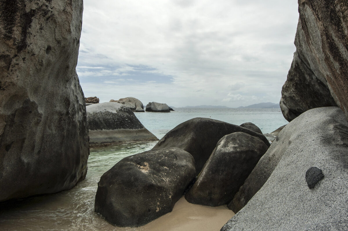 Large rounded granite boulders at The Baths on Virgin Gorda forming a natural passage to the sea, with shallow water and distant islands under a partly cloudy sky.