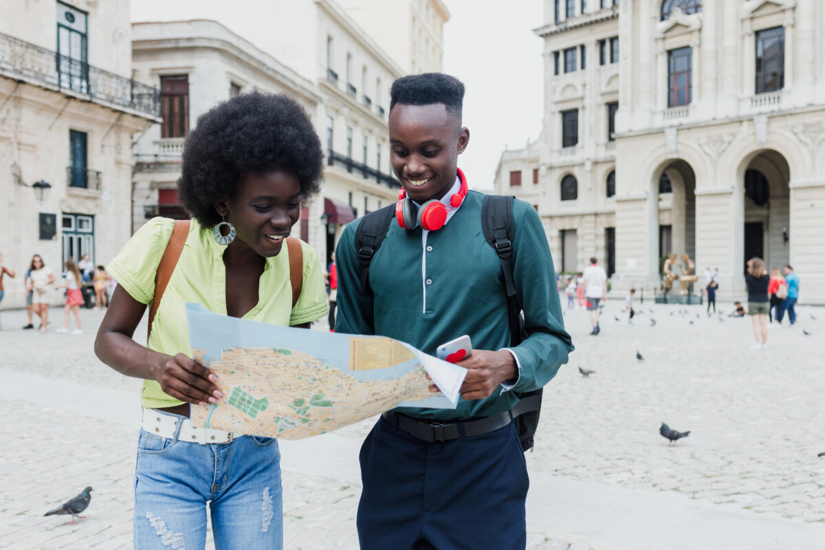 Two smiling travelers stand in a cobblestone plaza holding a city map, surrounded by historic stone buildings.