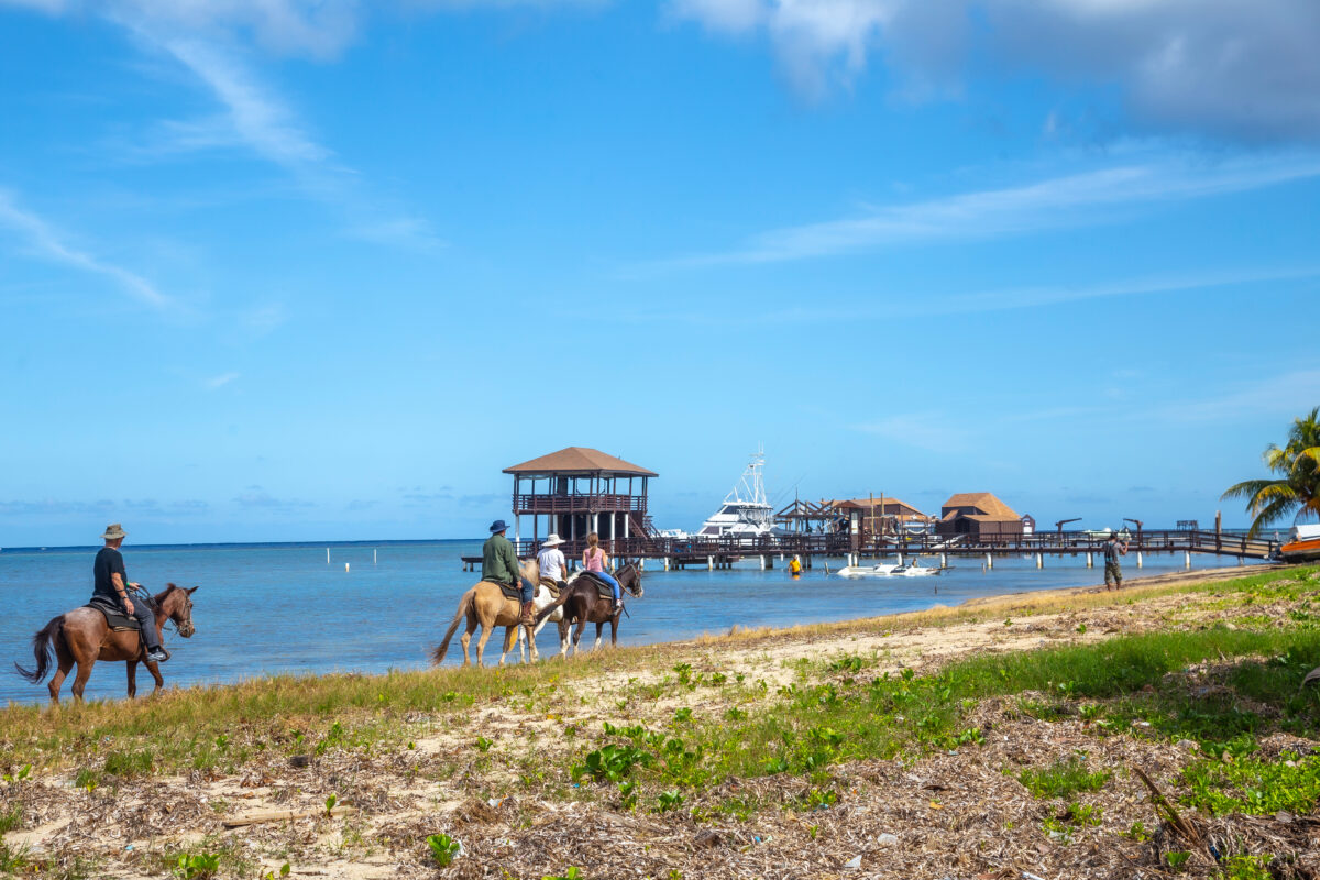 Several people on horseback riding along a sandy beach with wooden pavilions, moored boats, and turquoise water in Roatán, Honduras.
