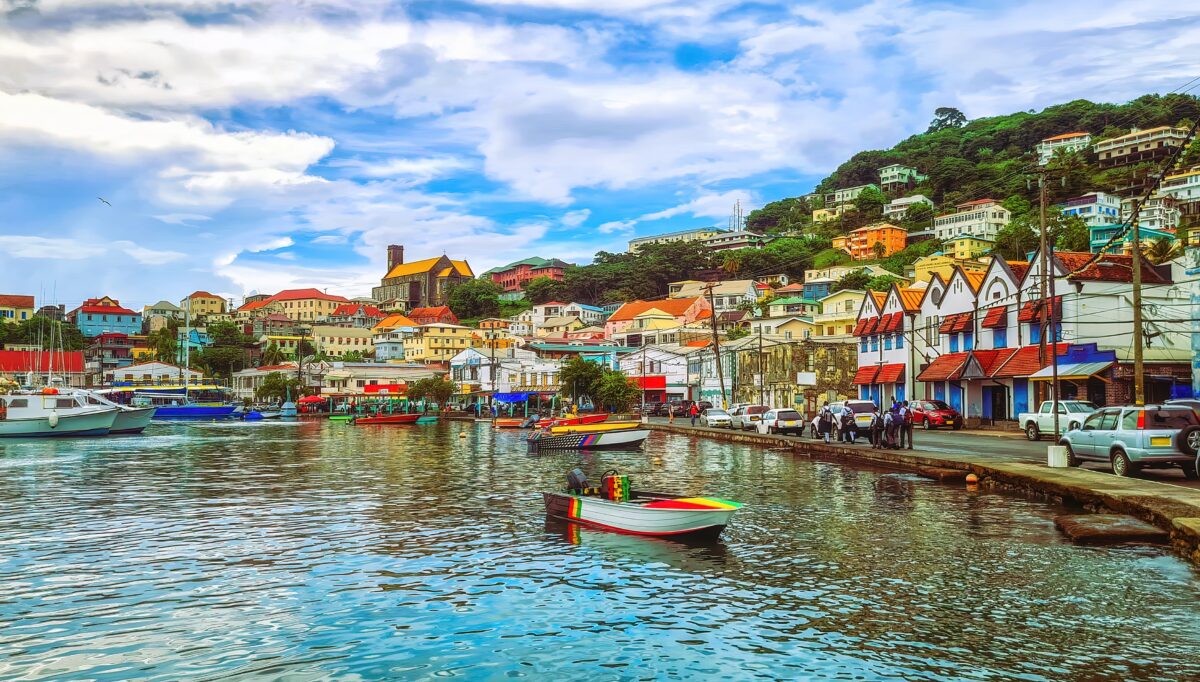 Eastern vs Western vs Southern Caribbean | Colorful hillside view of St. George’s, Grenada, showing rows of pastel buildings with red tile roofs rising above a harbor dotted with small fishing boats and calm, reflective water under a partly cloudy blue sky.
