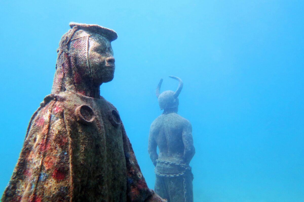 Two life-sized underwater statues in Grenada’s Underwater Sculpture Park, one facing forward with textured coral growth and another with horns seen from behind in clear blue water.