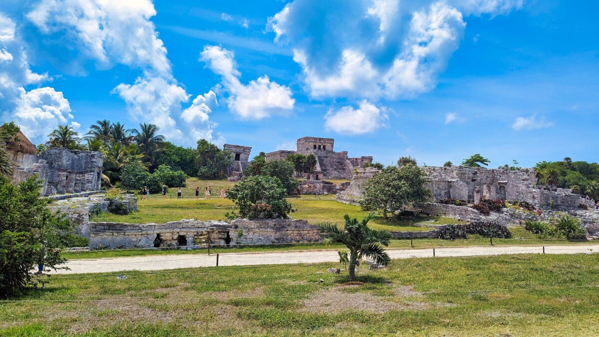 Ancient Mayan ruins at Tulum, showing limestone buildings and temple remains atop a green hillside with visitors walking among them beneath sunny skies.