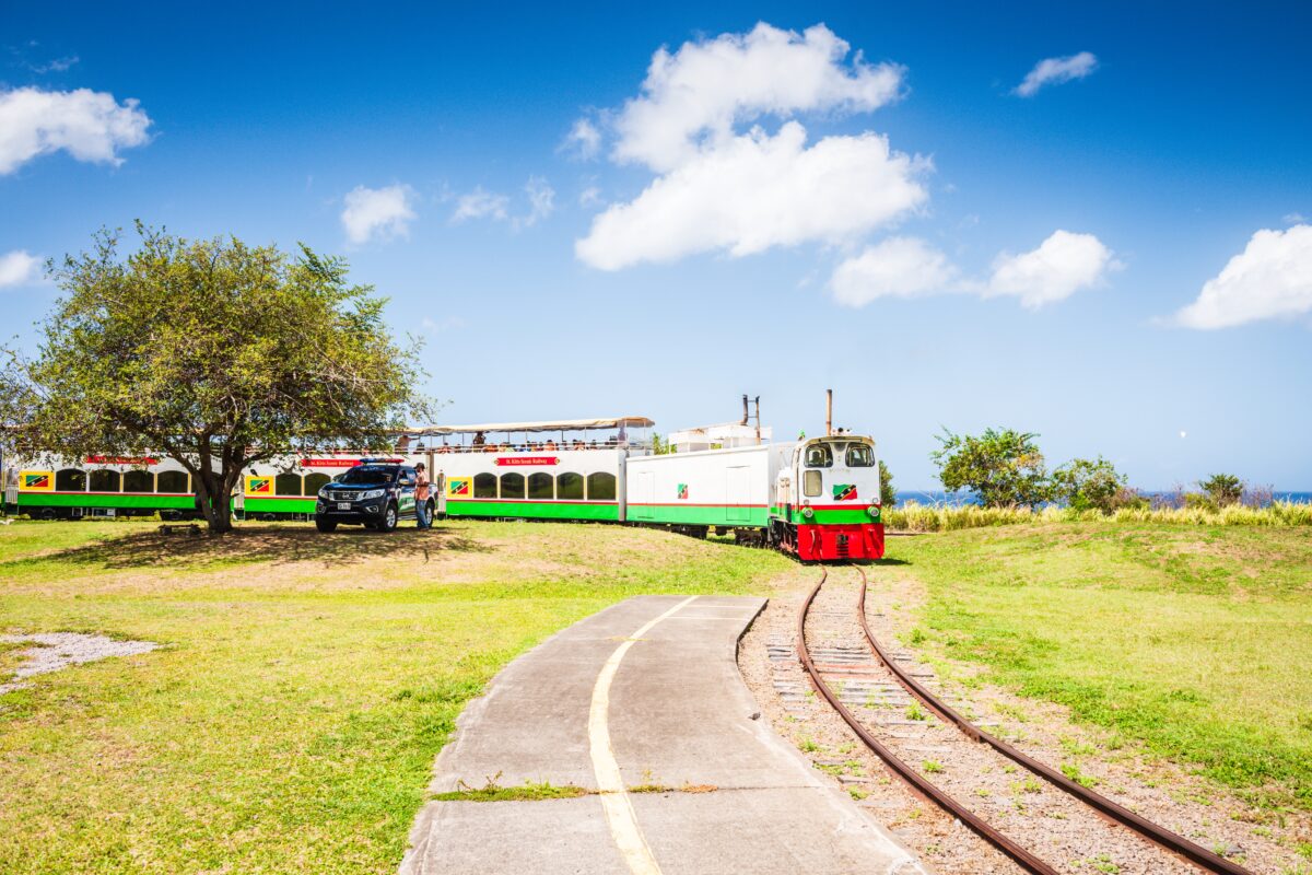 A bright green-and-white train with red trim travels along a narrow-gauge track under a blue sky. A single tree and parked vehicle stand nearby on grassy terrain.