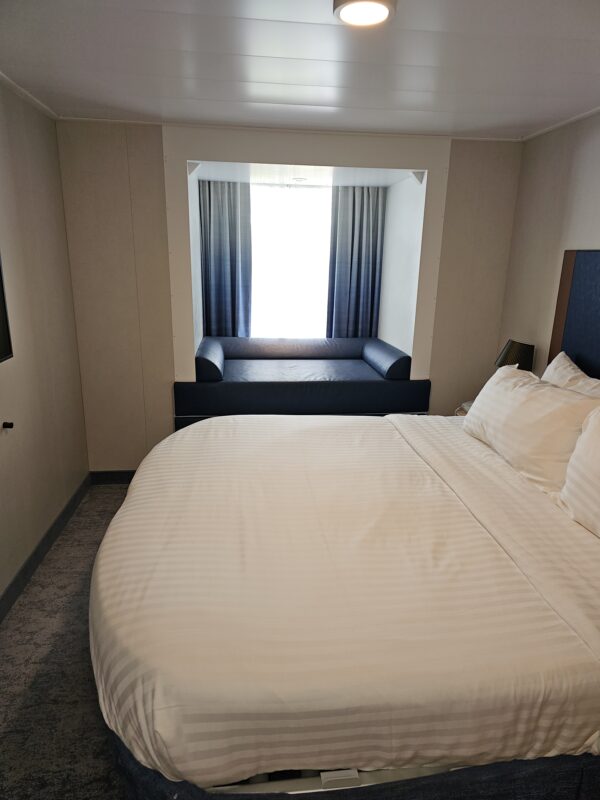 Cruise ship interior-view cabin with large window seat overlooking inside area of ship, showing bed and neutral décor.