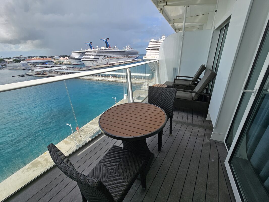Cruise ship balcony with chairs and table overlooking turquoise water and docked ships in port, showing spacious private outdoor area.