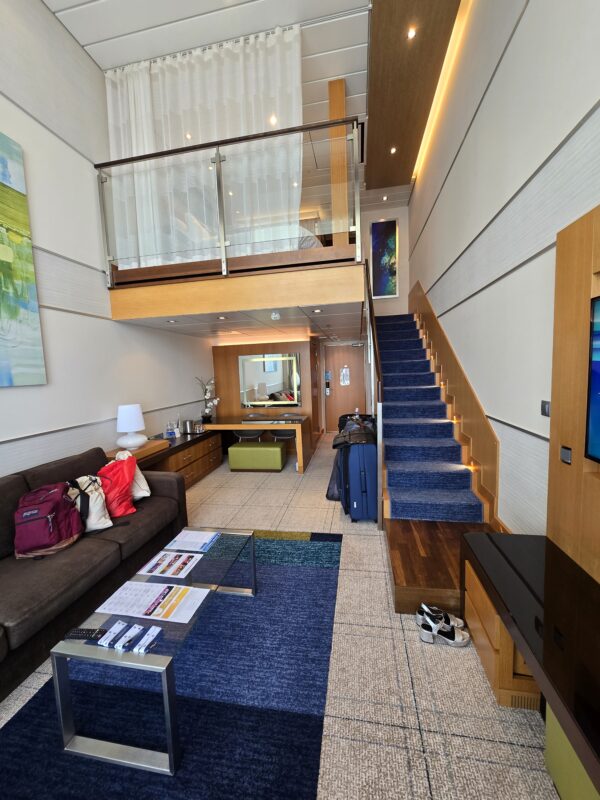 Two-level cruise ship suite with staircase, sofa, coffee table, and loft-style bedroom above, showing spacious modern layout.