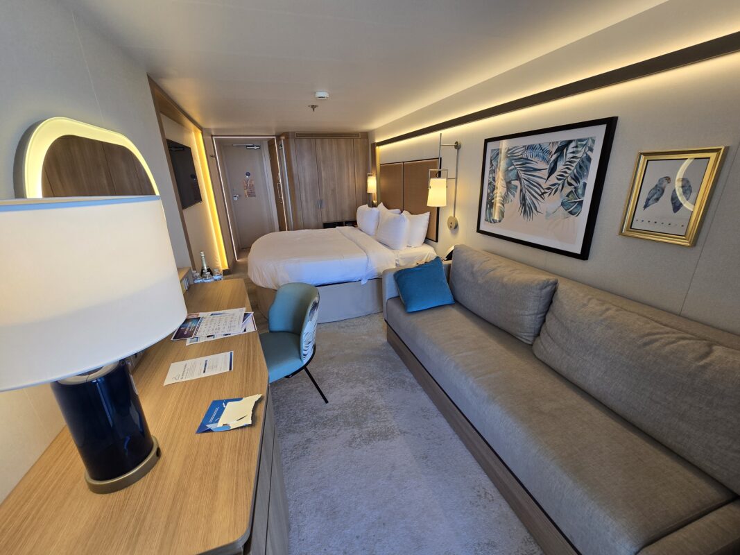 Contemporary cruise cabin interior featuring a double bed, long sofa, framed artwork, and desk area with lamp and chair.