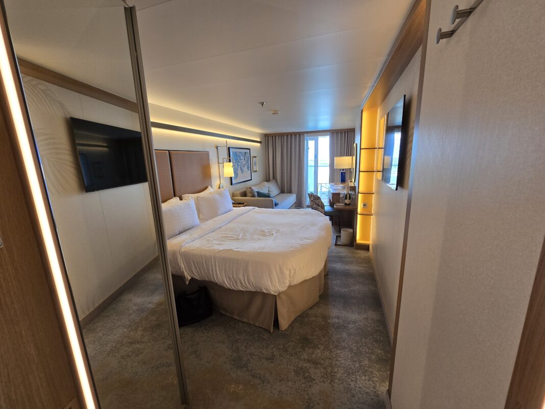 Modern cruise balcony stateroom with king bed, sofa, desk, and floor-to-ceiling glass doors opening to a private balcony.