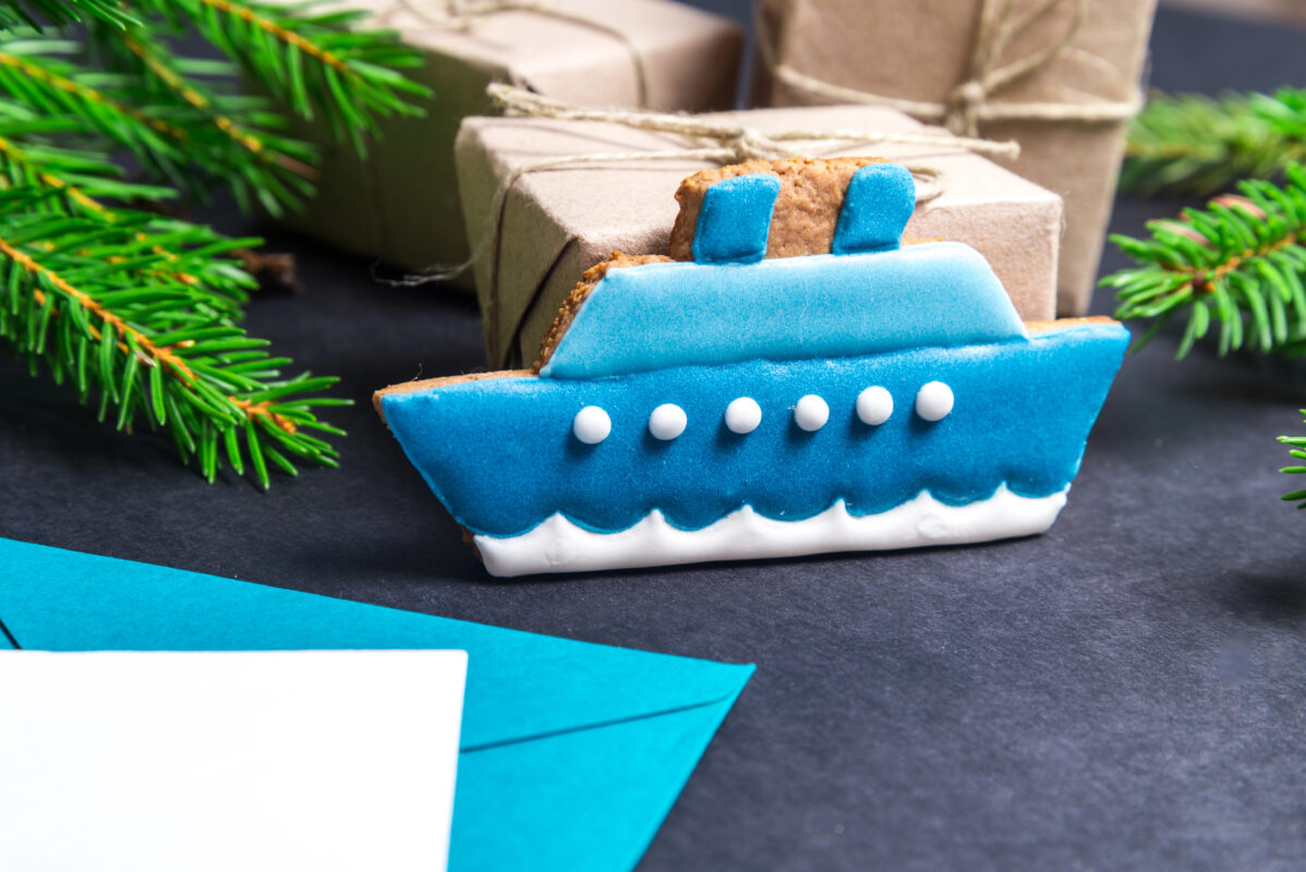 Close-up of a blue cruise-ship-shaped Christmas cookie propped against small wrapped gifts with evergreen branches around it.