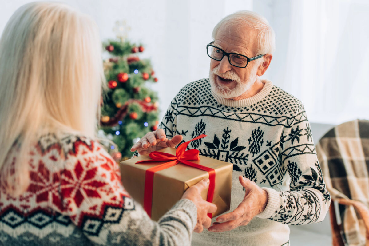 Close-up of an elderly man wearing glasses and a patterned sweater, accepting a Christmas present from a woman with long blonde hair.