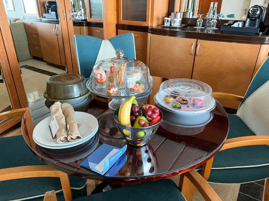 Table inside a Royal Caribbean suite displaying covered trays with desserts, shrimp appetizers, fruit, and bottled water—part of a Royal Up suite welcome.