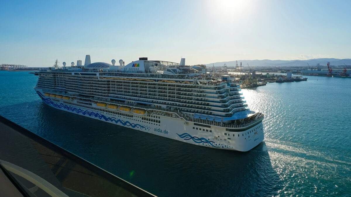 A large AIDA cruise ship sailing out of port, viewed from above, with multiple passenger decks and a wide white hull reflecting in the water.