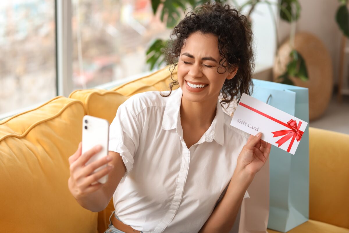 Smiling woman sitting on a yellow sofa, holding a white gift card envelope with a red ribbon while taking a selfie on her phone.