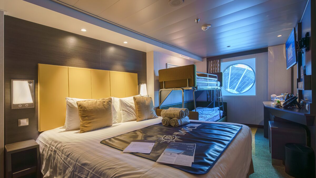 Family-friendly cruise cabin featuring a large bed, stacked bunks, and oceanview porthole, illustrating efficient design for four guests.
