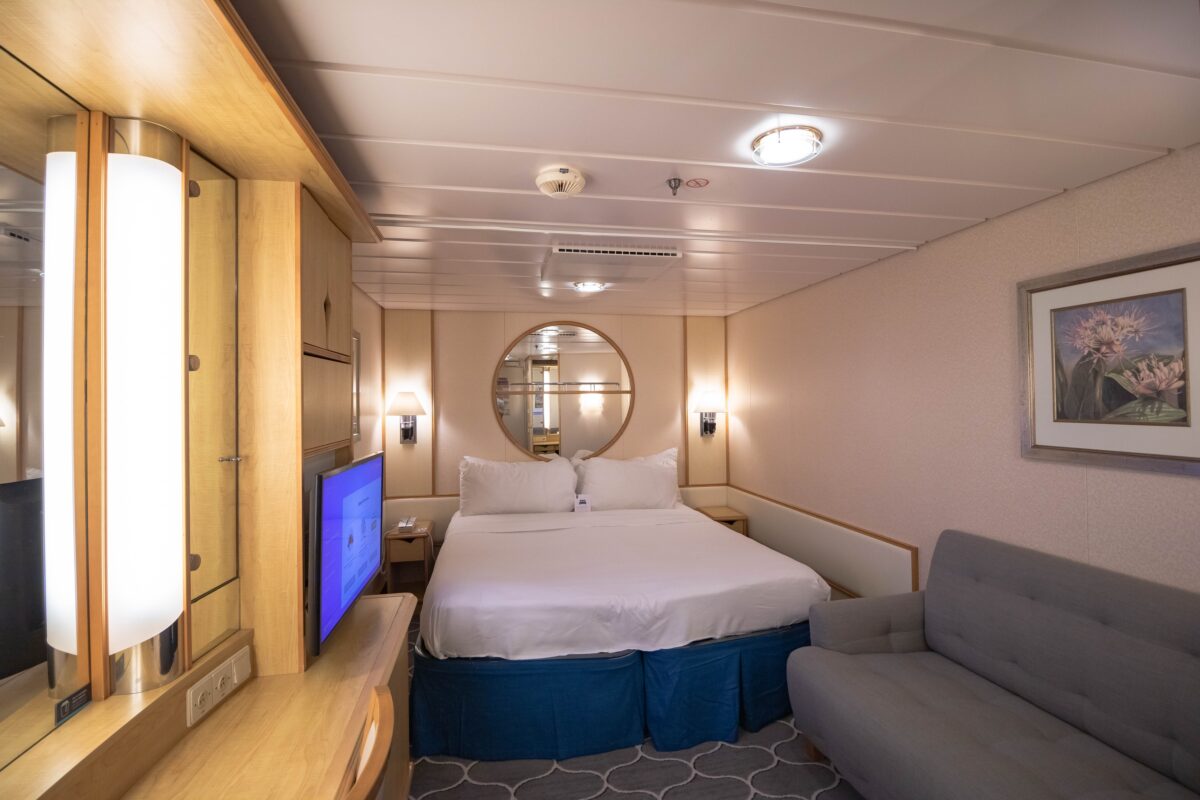 Choosing a Cruise Cabin |Interior cruise ship cabin with queen bed, sofa, and mirror on wall, showing a compact, well-lit layout without windows.
