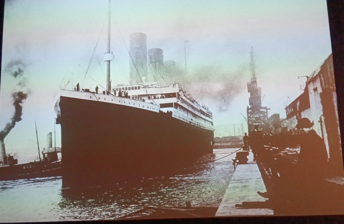 Vintage photo of a large ocean liner docked at a pier, viewed from the front, with multiple smokestacks and a tugboat alongside.